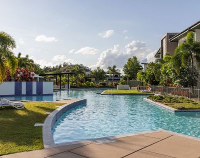 A serene outdoor pool surrounded by lush greenery and palm trees under a clear blue sky, conveying a peaceful, inviting, and tropical atmosphere.