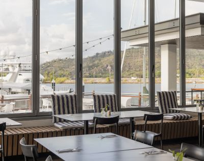 Modern restaurant interior with large windows showcasing a marina view. Tables set simply, striped cushions add comfort, creating a relaxed, inviting atmosphere.