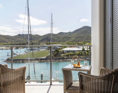 Two wicker chairs and a small table with drinks on a balcony overlook a serene marina with sailboats, lush green hills, and a clear blue sky. Peaceful ambiance.