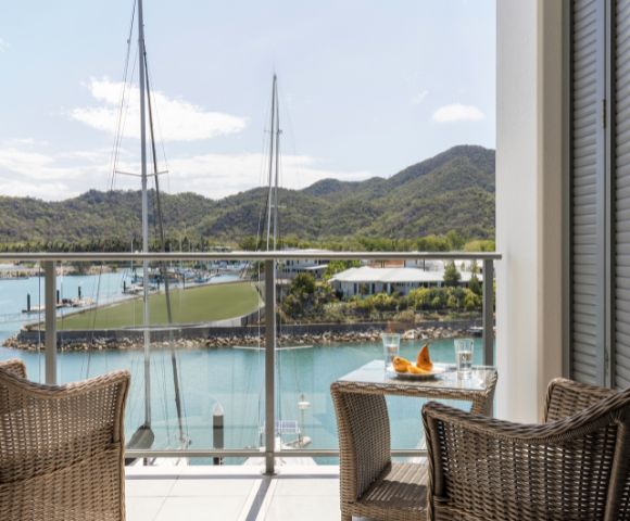 Two wicker chairs and a small table with drinks on a balcony overlook a serene marina with sailboats, lush green hills, and a clear blue sky. Peaceful ambiance.
