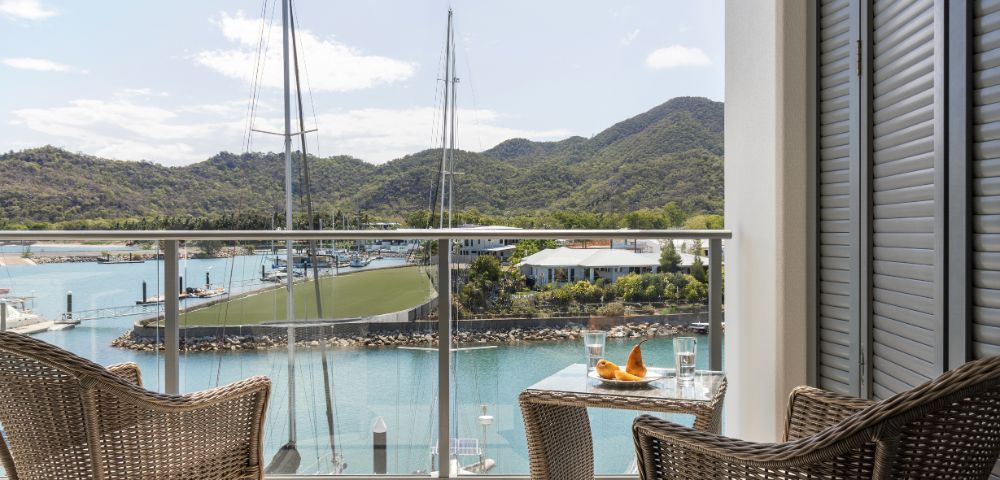 Two wicker chairs and a small table with drinks on a balcony overlook a serene marina with sailboats, lush green hills, and a clear blue sky. Peaceful ambiance.