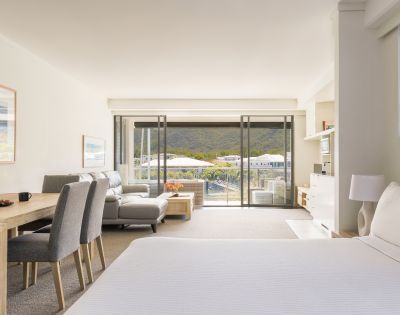 A bright, airy hotel room with a double bed, seating area, and a dining table. Large windows open to a scenic mountain view, conveying calm and relaxation.