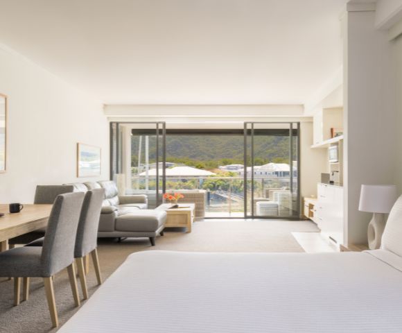 A bright, airy hotel room with a double bed, seating area, and a dining table. Large windows open to a scenic mountain view, conveying calm and relaxation.