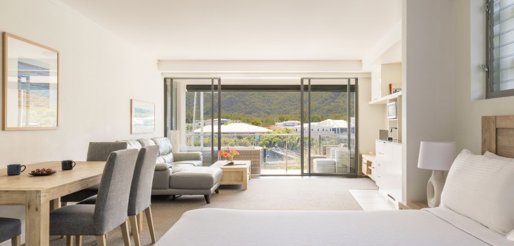 A bright, airy hotel room with a double bed, seating area, and a dining table. Large windows open to a scenic mountain view, conveying calm and relaxation.