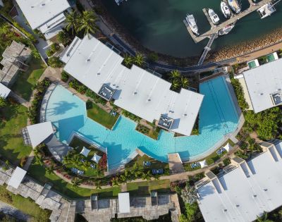 Aerial view of a luxurious, winding blue swimming pool surrounded by modern white-roofed buildings, lush greenery, and a marina with docked boats.
