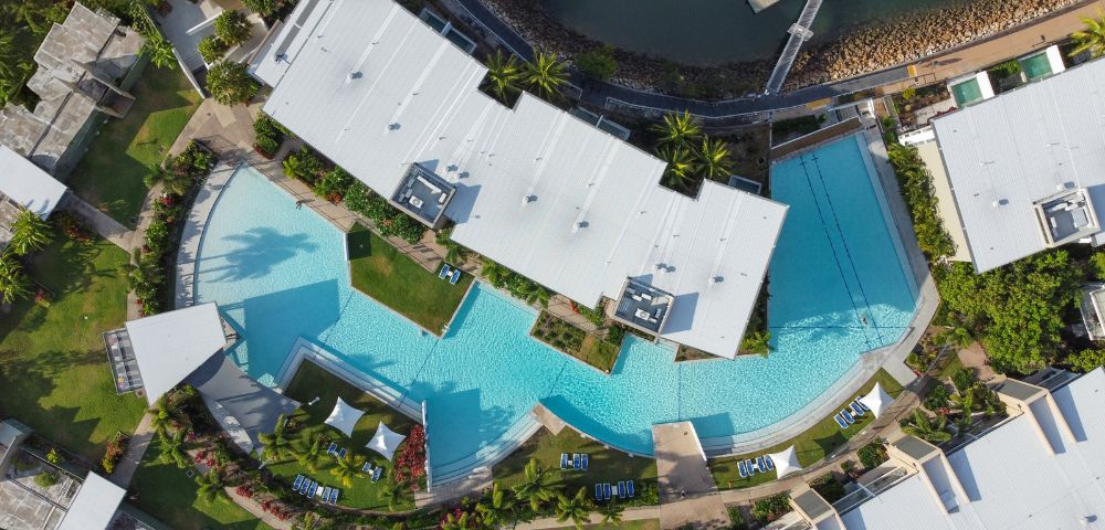Aerial view of a luxurious, winding blue swimming pool surrounded by modern white-roofed buildings, lush greenery, and a marina with docked boats.