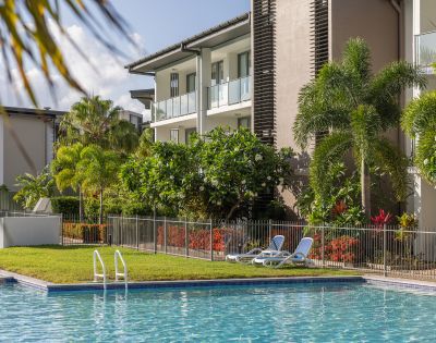 A serene outdoor pool area with reclined chairs on grass, surrounded by lush tropical plants and trees. Modern apartment building in the background under a clear sky.