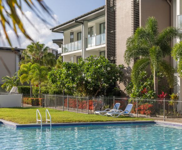 A serene outdoor pool area with reclined chairs on grass, surrounded by lush tropical plants and trees. Modern apartment building in the background under a clear sky.