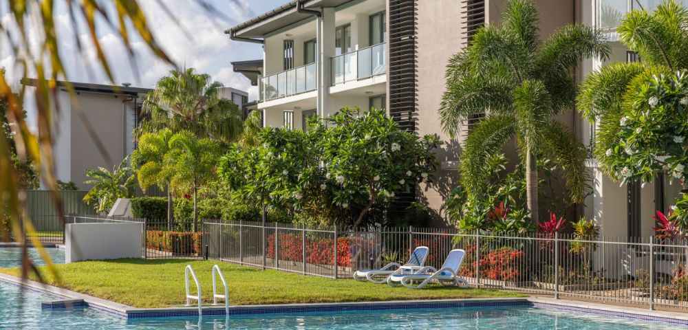 A serene outdoor pool area with reclined chairs on grass, surrounded by lush tropical plants and trees. Modern apartment building in the background under a clear sky.