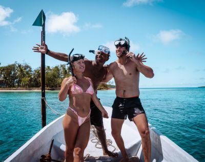 Three people in swimwear and snorkel gear pose joyfully on a boat against a backdrop of turquoise ocean and clear blue skies on a sunny day.