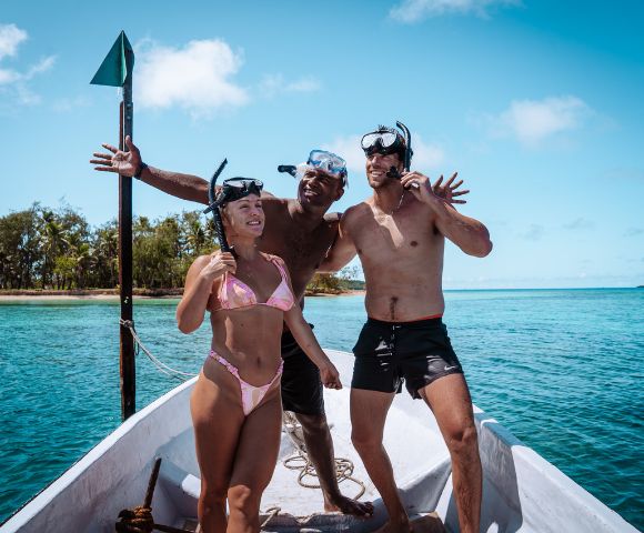 Three people in swimwear and snorkel gear pose joyfully on a boat against a backdrop of turquoise ocean and clear blue skies on a sunny day.
