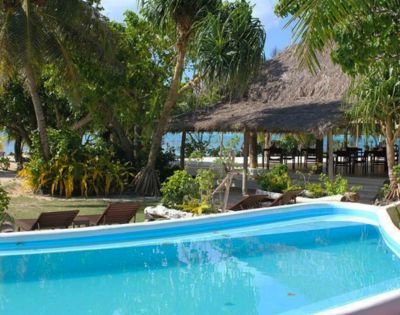 A tranquil tropical resort scene featuring a clear blue pool surrounded by lush greenery and palm trees, with a thatched-roof building and ocean view in the background.