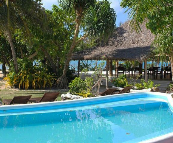 A tranquil tropical resort scene featuring a clear blue pool surrounded by lush greenery and palm trees, with a thatched-roof building and ocean view in the background.