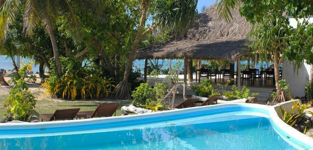 A tranquil tropical resort scene featuring a clear blue pool surrounded by lush greenery and palm trees, with a thatched-roof building and ocean view in the background.