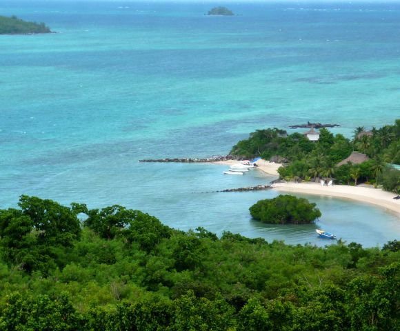 Scenic view of a tropical beach with turquoise waters, lush green vegetation, and a sandy shore. Boats are docked by the shore, creating a serene atmosphere.