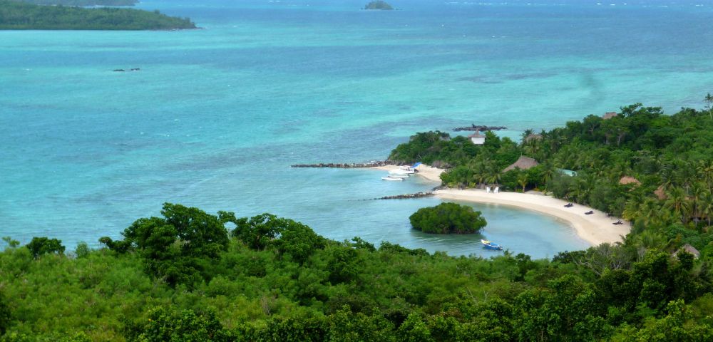 Scenic view of a tropical beach with turquoise waters, lush green vegetation, and a sandy shore. Boats are docked by the shore, creating a serene atmosphere.