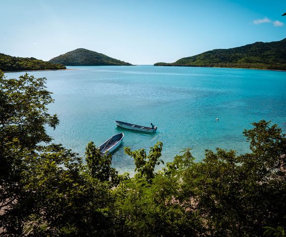A tranquil, clear blue sea framed by lush green hills under a bright sky. Two small boats float near the shore, conveying calm and serenity.