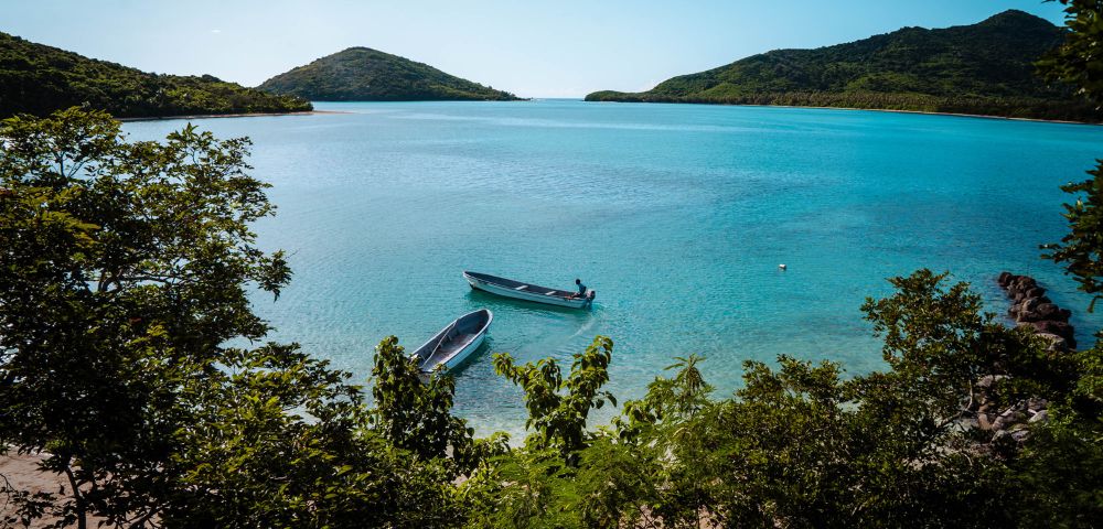 A tranquil, clear blue sea framed by lush green hills under a bright sky. Two small boats float near the shore, conveying calm and serenity.