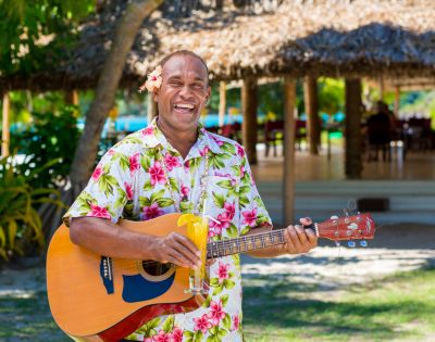 Man joyfully playing guitar, wearing a floral shirt with a flower behind his ear, holding a tropical drink. Sunny outdoor setting with a thatched hut background.