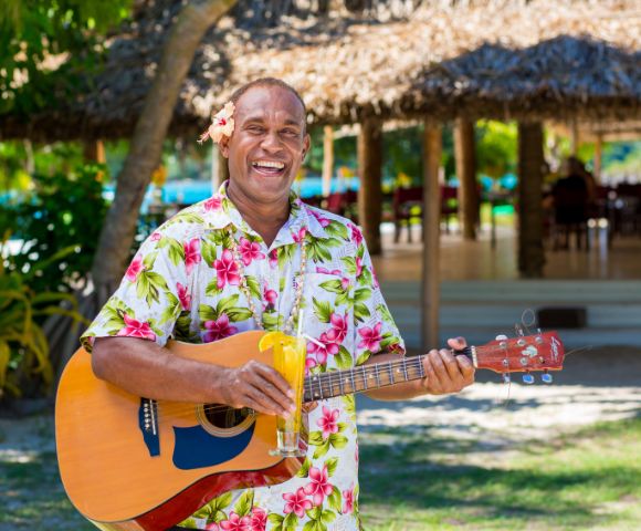 Man joyfully playing guitar, wearing a floral shirt with a flower behind his ear, holding a tropical drink. Sunny outdoor setting with a thatched hut background.