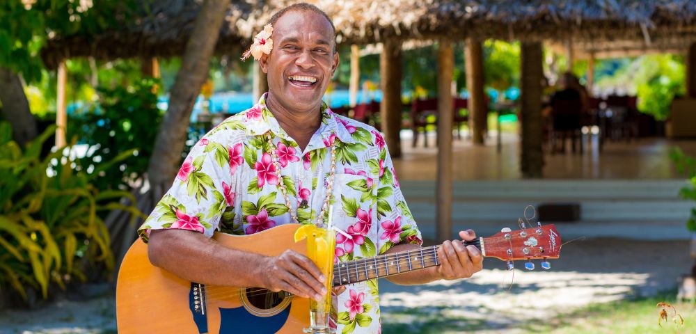 Man joyfully playing guitar, wearing a floral shirt with a flower behind his ear, holding a tropical drink. Sunny outdoor setting with a thatched hut background.