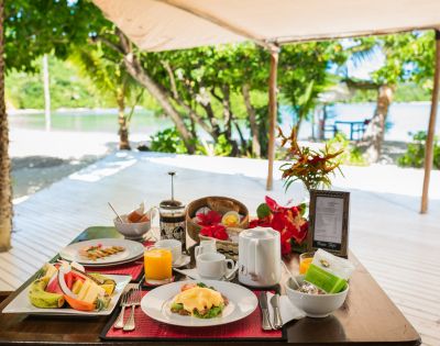 Outdoor table set for breakfast under a canopy near a tropical beach, featuring plates of fruit and eggs Benedict, a teapot, and vibrant flowers.