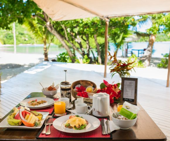 Outdoor table set for breakfast under a canopy near a tropical beach, featuring plates of fruit and eggs Benedict, a teapot, and vibrant flowers.