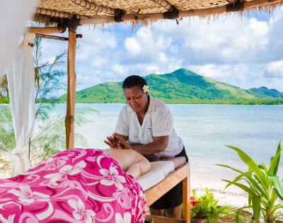 A serene beachside setting with a massage therapist giving a massage to a person covered in a pink floral towel. There's a view of lush green hills and clear blue water in the background.