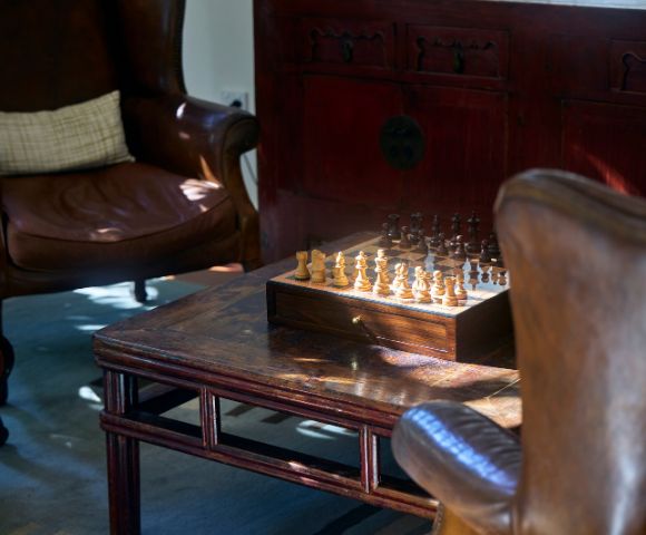 Chessboard on a wooden table between two leather armchairs in a cozy, warmly lit room. Sunlight casts gentle shadows, creating a peaceful ambiance.
