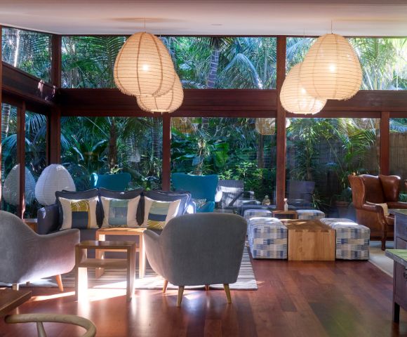 Modern living room with large windows showing lush greenery. Features cozy gray chairs, wooden floors, and soft lighting from pendant lamps, creating a serene atmosphere.