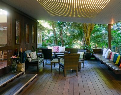 Covered patio with wooden flooring and tropical plants. Features cozy seating with colorful cushions, creating a serene, inviting atmosphere.