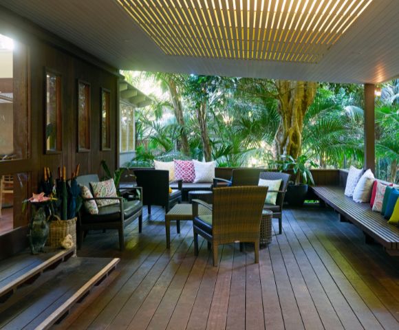 Covered patio with wooden flooring and tropical plants. Features cozy seating with colorful cushions, creating a serene, inviting atmosphere.