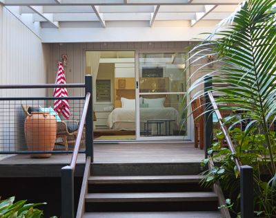 Cozy patio leading to a serene bedroom with open sliding glass doors. Lush greenery surrounds the deck and a striped towel hangs near a large vase.