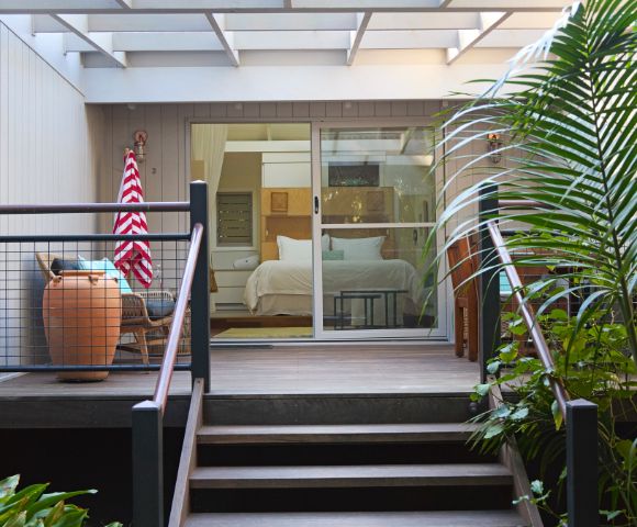 Cozy patio leading to a serene bedroom with open sliding glass doors. Lush greenery surrounds the deck and a striped towel hangs near a large vase.