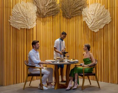 A couple dines at a round table in a stylish restaurant with wood-paneled walls and large decorative fans. A waiter serves wine, creating an intimate, elegant atmosphere.