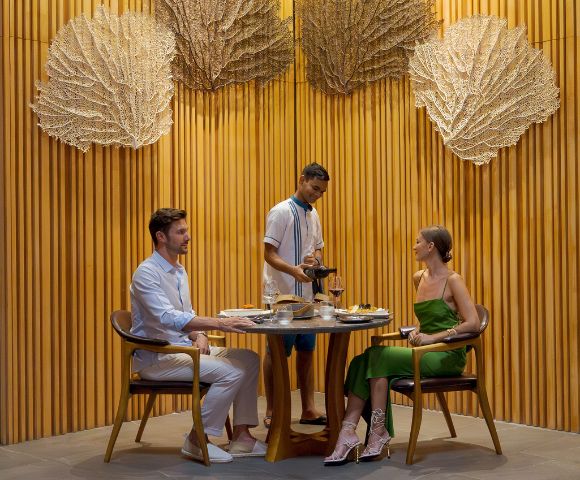 A couple dines at a round table in a stylish restaurant with wood-paneled walls and large decorative fans. A waiter serves wine, creating an intimate, elegant atmosphere.
