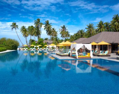Luxurious tropical resort with a large blue pool, surrounded by palm trees, yellow sun umbrellas, and cabanas under a clear blue sky, conveying relaxation.