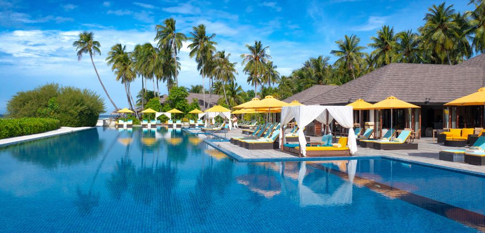 Luxurious tropical resort with a large blue pool, surrounded by palm trees, yellow sun umbrellas, and cabanas under a clear blue sky, conveying relaxation.