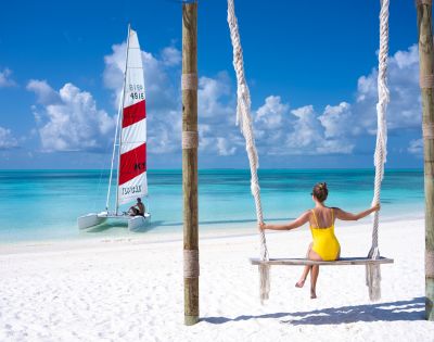A woman in a yellow swimsuit sits on a swing facing a turquoise sea. A sailboat is on the water under a blue sky with fluffy clouds, creating a serene scene.