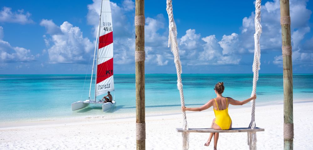 A woman in a yellow swimsuit sits on a swing facing a turquoise sea. A sailboat is on the water under a blue sky with fluffy clouds, creating a serene scene.