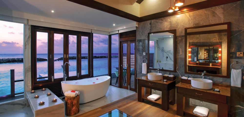 Luxurious bathroom with ocean views, a freestanding tub, and double sinks on wooden vanities. Warm lighting and candles create a serene atmosphere.