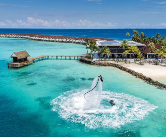 A person on a water jetpack high above turquoise ocean waters, near a tropical resort with overwater bungalows and palm trees, under a clear blue sky.
