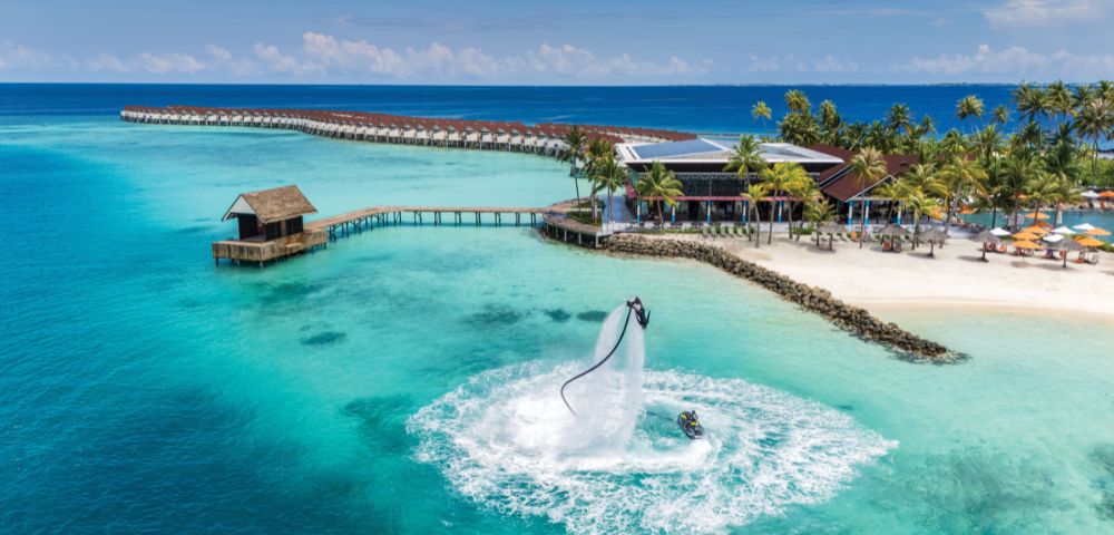A person on a water jetpack high above turquoise ocean waters, near a tropical resort with overwater bungalows and palm trees, under a clear blue sky.