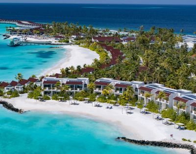 Aerial view of a tropical resort with villas along a pristine white sand beach, turquoise waters, and lush green palm trees under a clear blue sky.