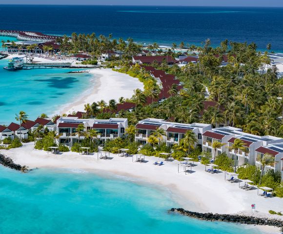 Aerial view of a tropical resort with villas along a pristine white sand beach, turquoise waters, and lush green palm trees under a clear blue sky.