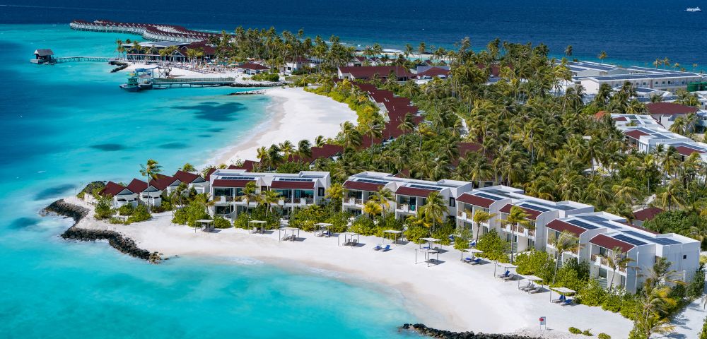 Aerial view of a tropical resort with villas along a pristine white sand beach, turquoise waters, and lush green palm trees under a clear blue sky.