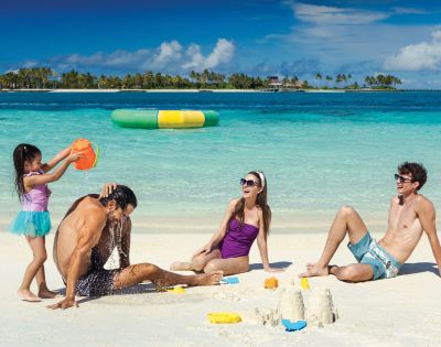 A family enjoys a beach day; a girl pours water on a man, a woman in a purple swimsuit sits smiling, and a man in swim trunks relaxes beside sandcastles.