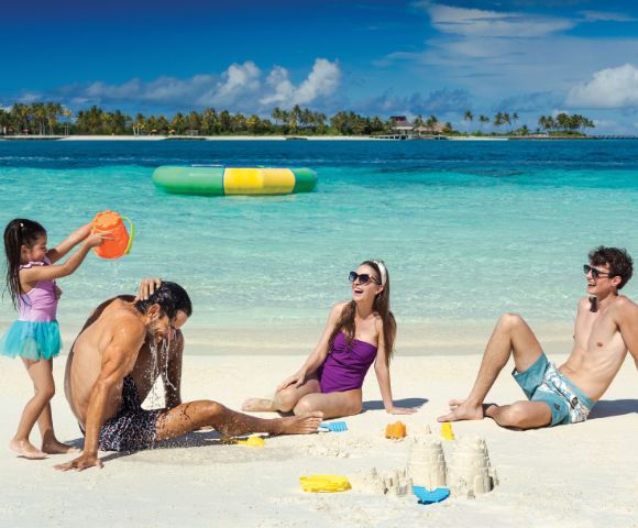 A family enjoys a beach day; a girl pours water on a man, a woman in a purple swimsuit sits smiling, and a man in swim trunks relaxes beside sandcastles.
