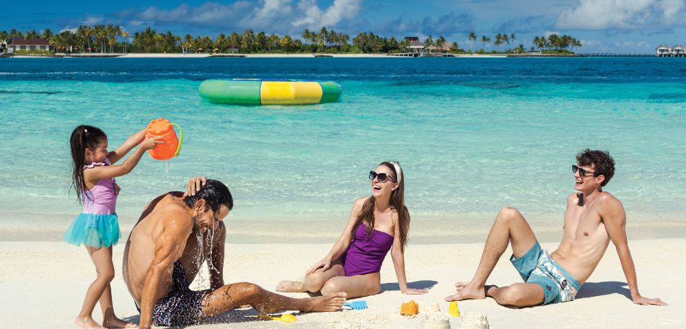 A family enjoys a beach day; a girl pours water on a man, a woman in a purple swimsuit sits smiling, and a man in swim trunks relaxes beside sandcastles.