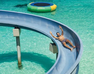 Man sliding down a blue waterslide into clear turquoise water, smiling and raising two fingers in a peace sign. Nearby, a colorful floating trampoline.